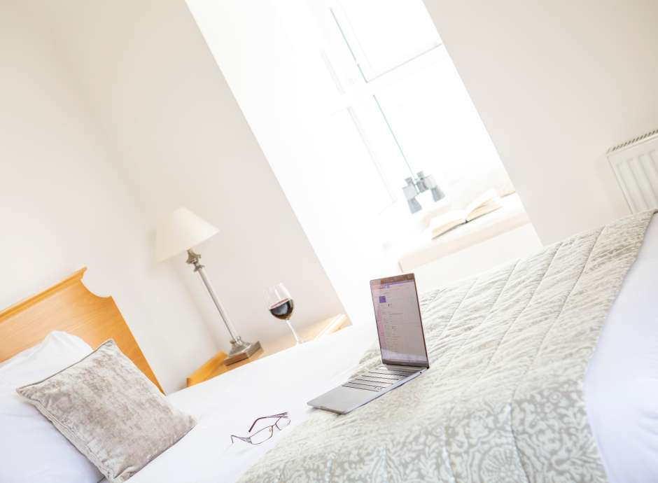 Laptop and glasses on a single hotel bed with light pouring in from large window