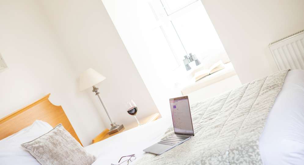 Laptop and glasses on a single hotel bed with light pouring in from large window