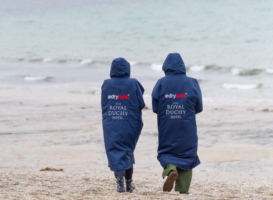 Couple walking along Falmouth beach on Autumn day wearing Royal Duchy Hotel branded dry robes